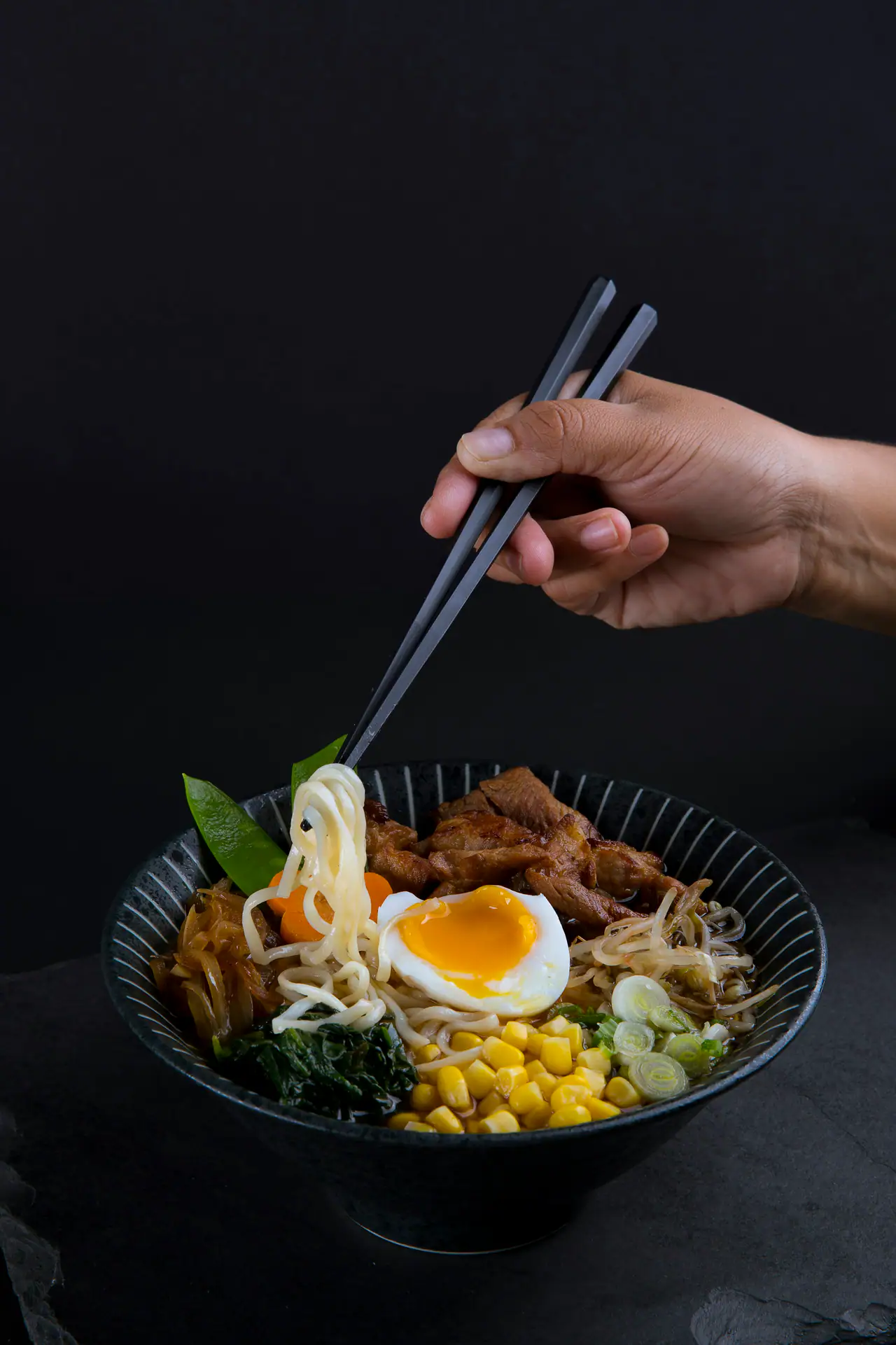 A hand lifts noodles with dark chopsticks from a black ramen bowl topped with a soft egg, corn, roast pork and scallions, against a dark backdrop