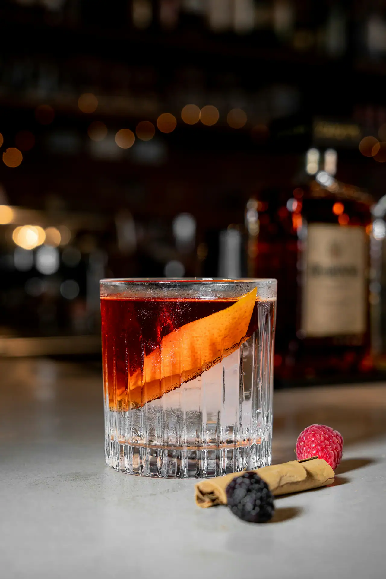 A stirred amber-red cocktail with a long orange peel in a cut-crystal glass on a pale bar top, with a raspberry and blackberry beside it and warm bokeh lights and bottles behind