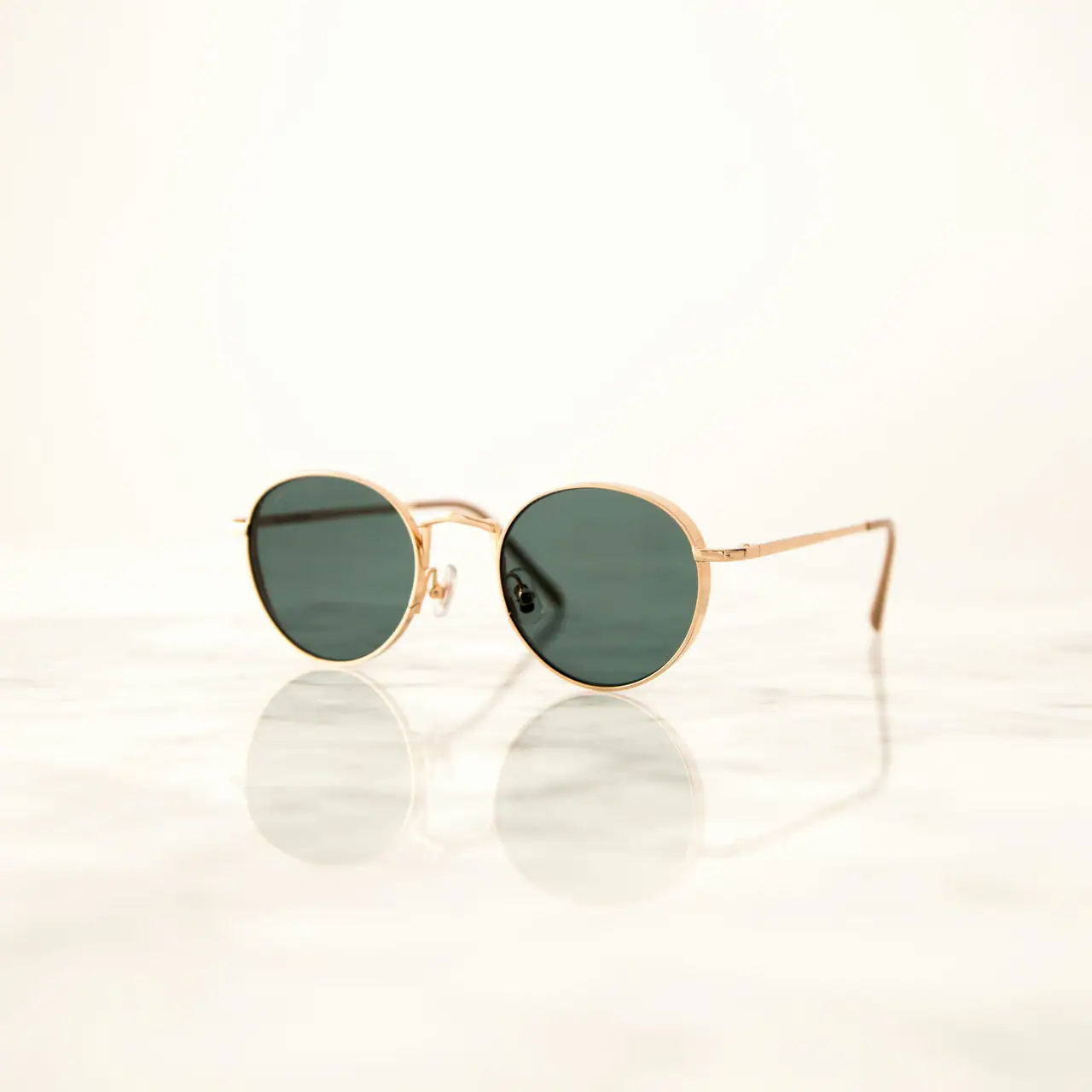 A pair of round gold-framed sunglasses with dark green lenses on a white marble surface, softly reflected