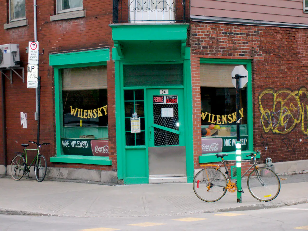 Corner storefront of Wilensky's Light Lunch on Avenue Fairmount in Mile End, with green trim, a red brick facade, hand-lettered yellow window signs and two bicycles at the curb