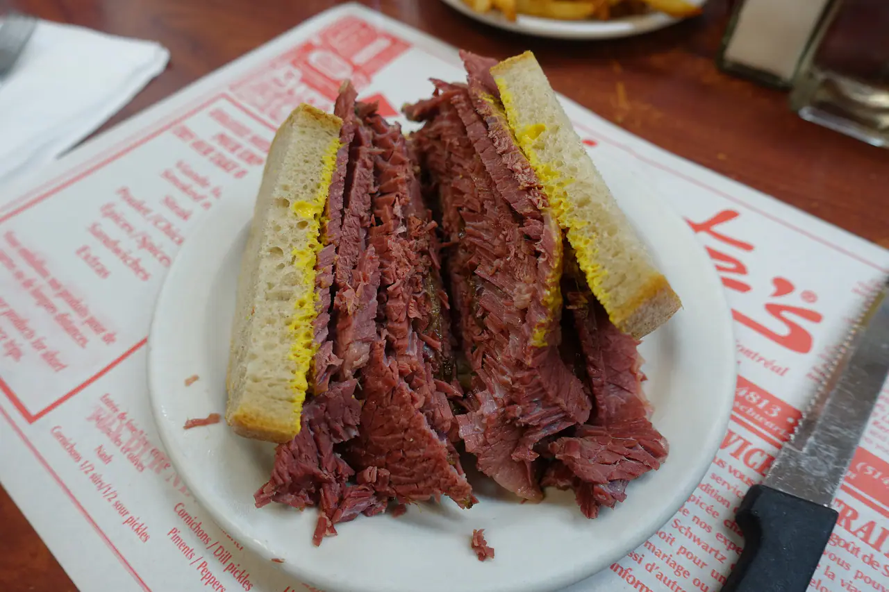 A tall Montréal smoked meat sandwich with yellow mustard, cut in half on a white plate at Schwartz's Deli in the Plateau, set on the deli's red-and-white printed placemat with a steak knife at right and a plate of fries behind