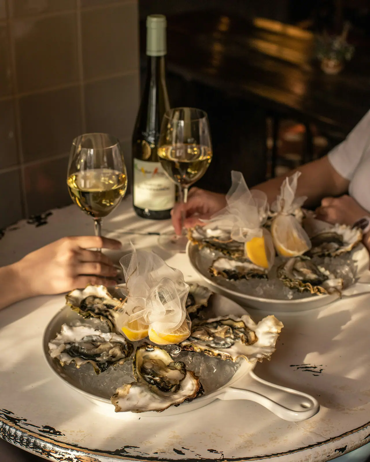 Two dishes of oysters on ice with lemon wrapped in tulle, two glasses of white wine and a bottle on a chipped white-painted table, two diners' hands at the table, lit by low golden sunlight against a dark background